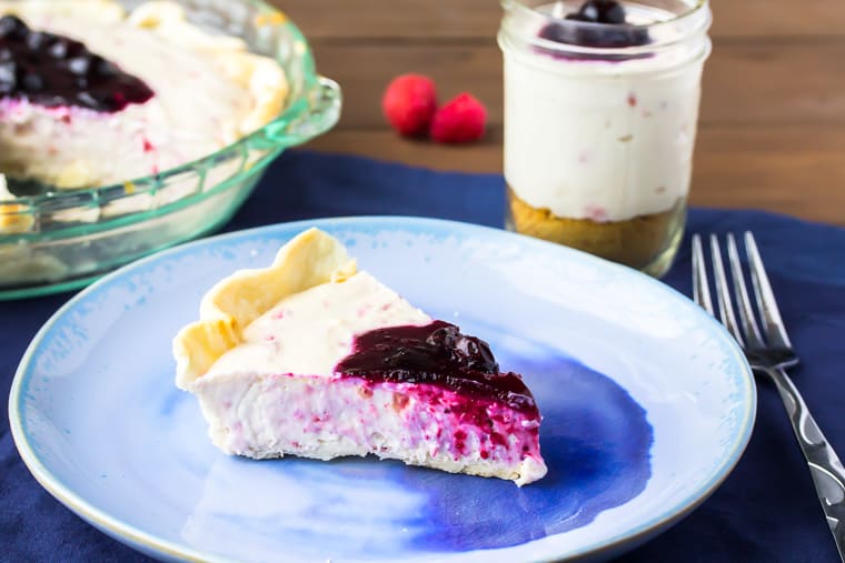 A slice of no bake raspberry cheesecake on a blue plate over a blue napkin with part of the whole pie showing and a mason jar filled with cheesecake, fork, and raspberries in the background