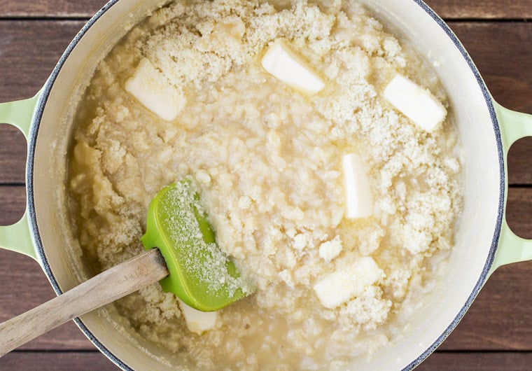 Butter and Parmesan Cheese added to the Risotto in a Dutch oven with a green and wood spatula in it over a wood background