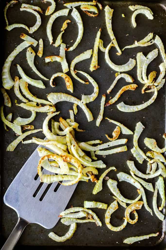 A sheet pan with baked fennel slices on it with a silver spatula scooping some up