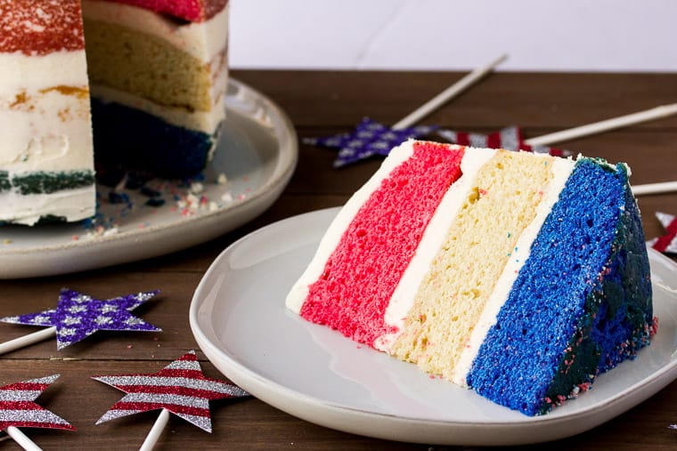 A Large Slice of Red, White, and Blue Cake on a White Plate with the Whole Cake and Patriotic Star Decorations in the back ground