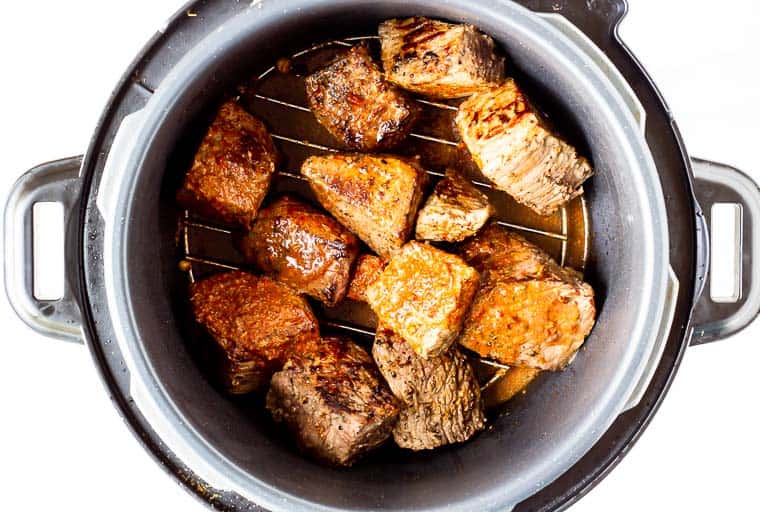 Beef cubes covered in marinade in the bottom of a pressure cooker over a white background