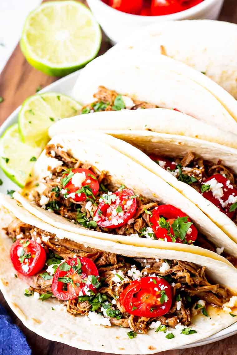 Close up of instant pot beef barbacoa tacos on a white plate over a wood table with a lime half and a white bowl of tomatoes in the background