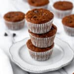 Three pumpkin chocolate chip muffins stacked on top of each other on a small white plate with more muffins scattered in the background.