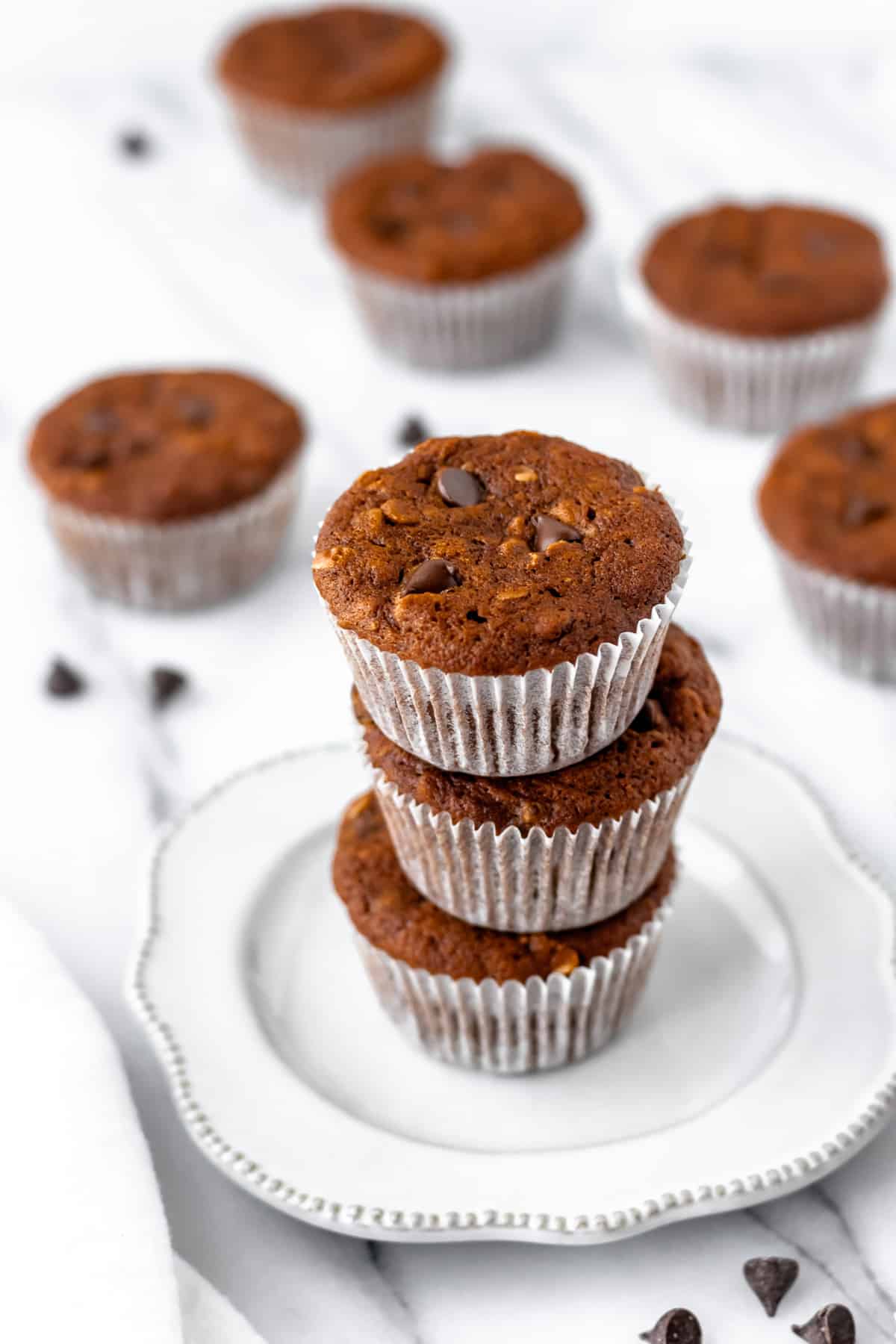 Three pumpkin chocolate chip muffins stacked on top of each other on a small white plate with more muffins scattered in the background.