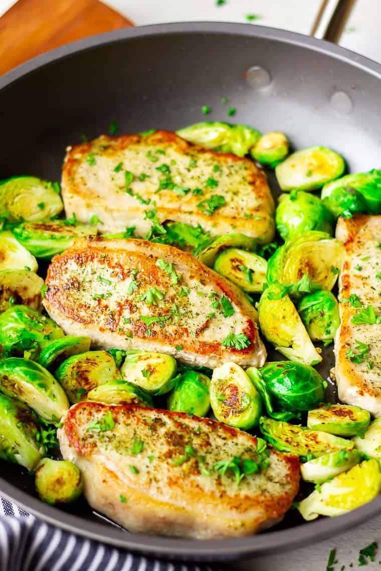 Close up of 4 Pan Fried Ranch Pork Chops and Brussels Sprouts in a black skillet with a wood spatula in the background