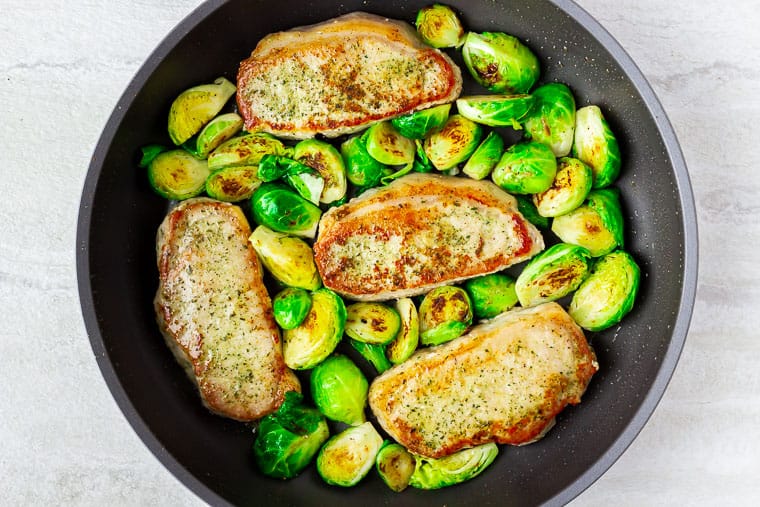 4 pork chops and brussels sprouts in a black skillet over a white background