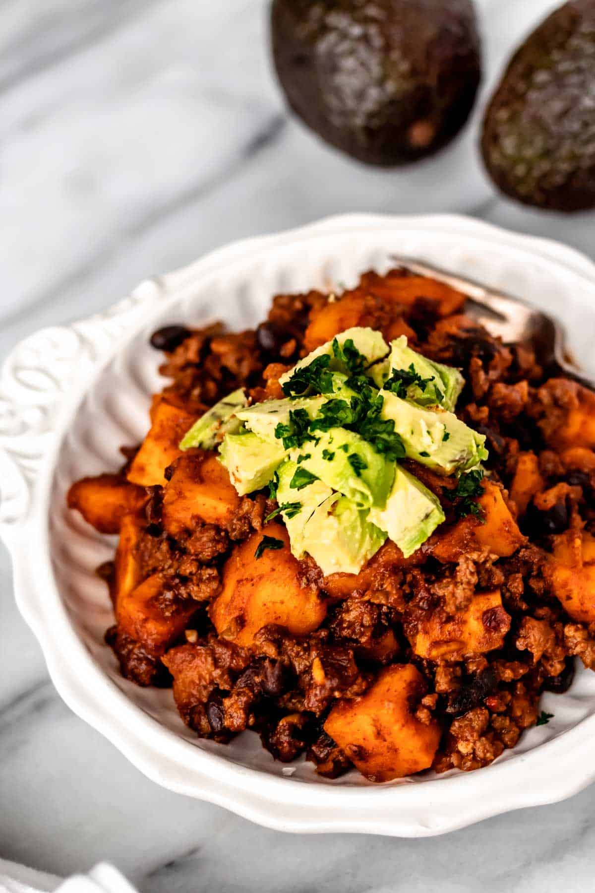Close up of a bowl of slow cooker sweet potato chili with 2 avocados in the background.