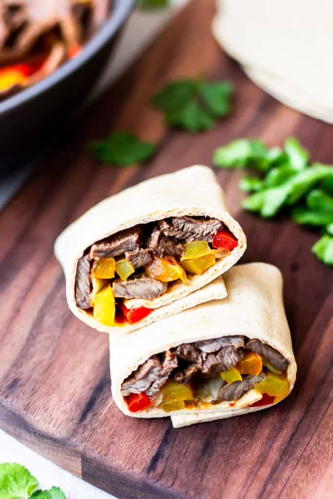Steak fajitas wrapped in tortillas on a wood board with part of a skillet with peppers and beef in it, cilantro leaves, and tortillas in the background