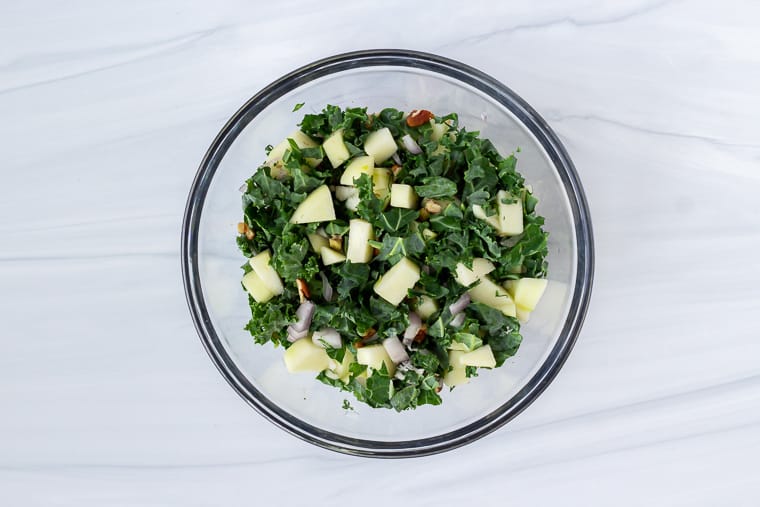 Chopped kale, apples, shallot, and pecans in a glass bowl over a white background