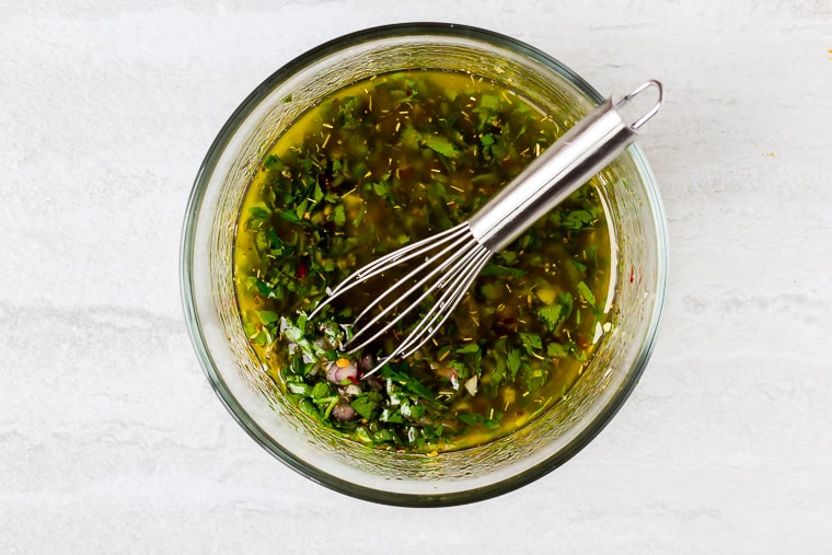 Chimichurri sauce in a glass bowl with a whisk over a white background