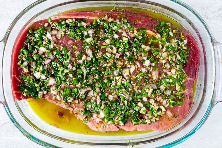 A large piece of steak marinating in chimichurri sauce in a glass baking dish over a white background