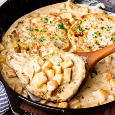 A pork chop topped with apples in a maple cream sauce being lifted out with a wood turner from a cast iron skillet that's sitting on a wood board over a white background with 2 apples and a blue and white striped towel near by