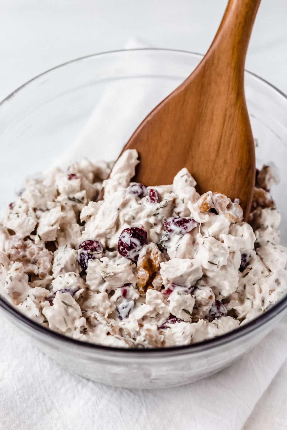 A glass bowl of cranberry chicken salad with a wood spoon in it over a white background