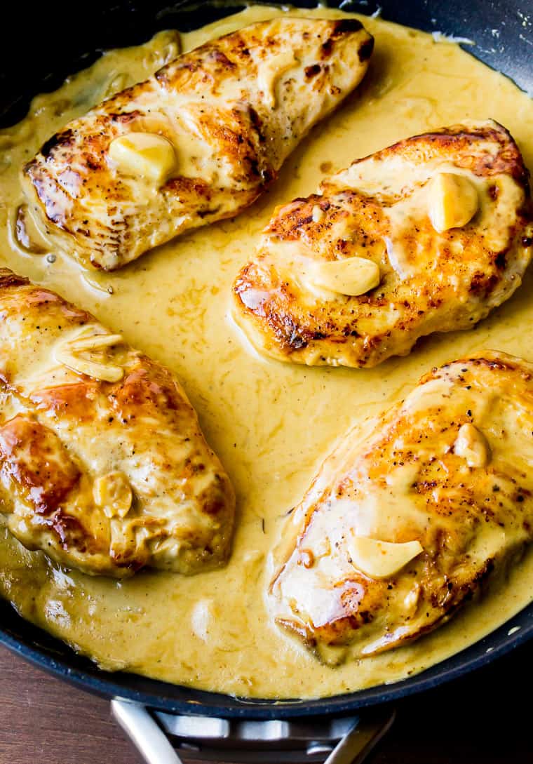 Close up of 4 chicken breasts in sauce, topped with creamy garlic sauce and slices of garlic in a black skillet