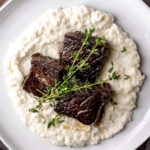 Overhead view of three short ribs on a bed of mashed cauliflower with thyme sprigs as garnish.
