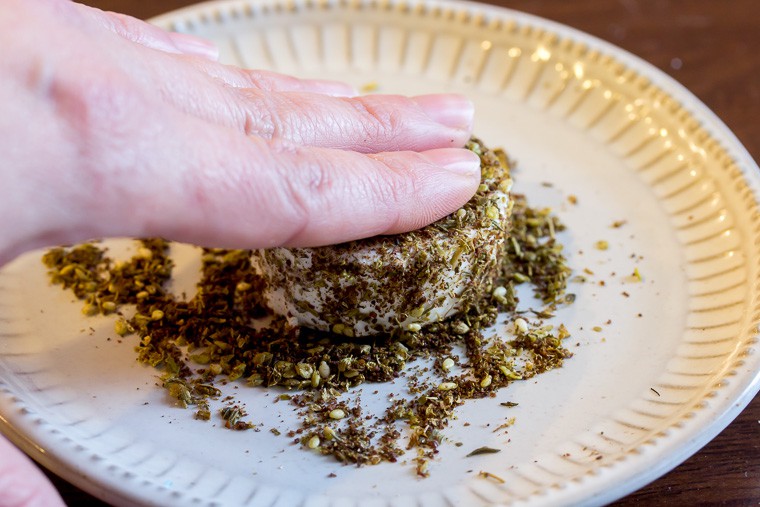 Close up of fingers pressing a round of goat cheese into za'atar spices on a white plate