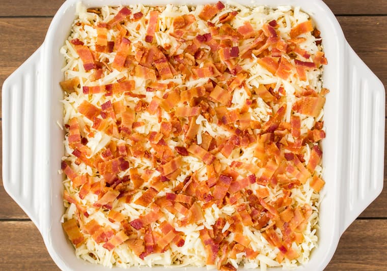 Close up overhead view of the Keto Breakfast Casserole prepared and ready to bake topped with bacon and more cheese in a white casserole dish on a wood background