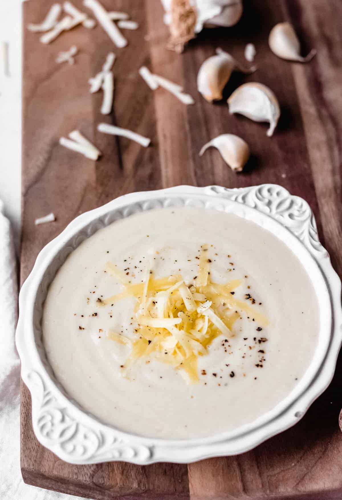 Close up of White cheddar cauliflower soup in a white bowl on a wood board with a head of garlic, garlic cloves, and white cheddar cheese