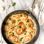 Creamy garlic chicken in a black skillet on a marble background with garlic cloves and parsley around it with text overlay.
