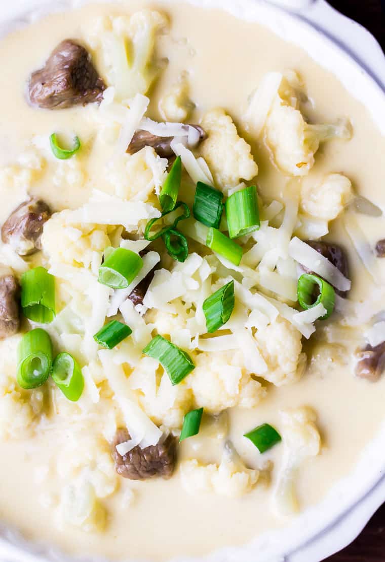 Close up of a bowl of beef and cauliflower soup topped with sliced green onions and shredded cheese