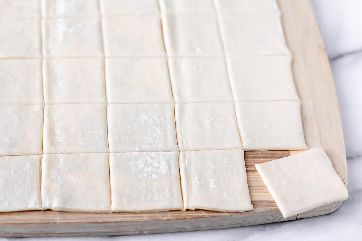 Puff pastry cut into small squares on a wood cutting board.