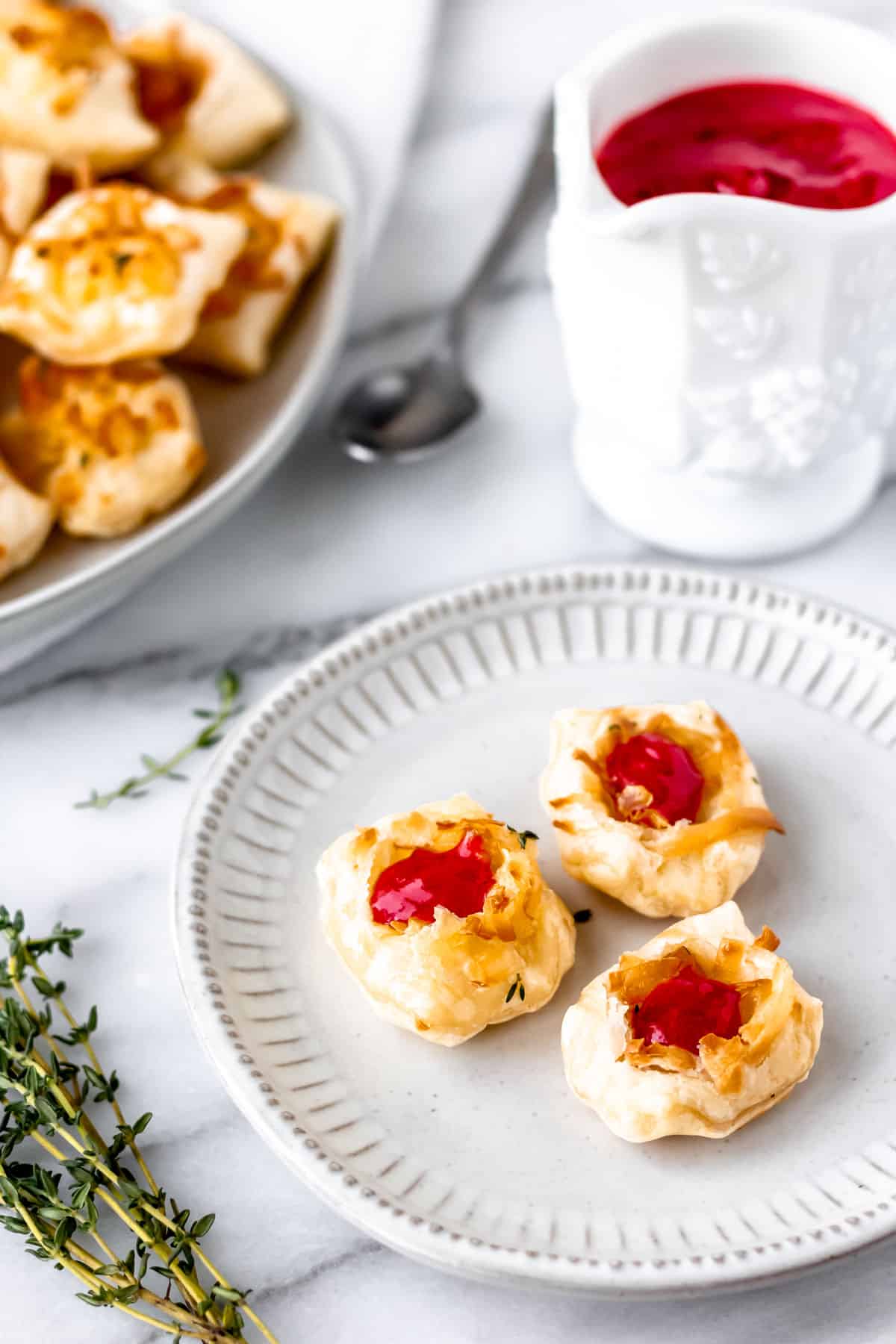 Three cheese puffs with raspberry sauce on a small white plate with a second plate of Gruyere cheese puffs, a pitcher of raspberry sauce and a spoon in the background.