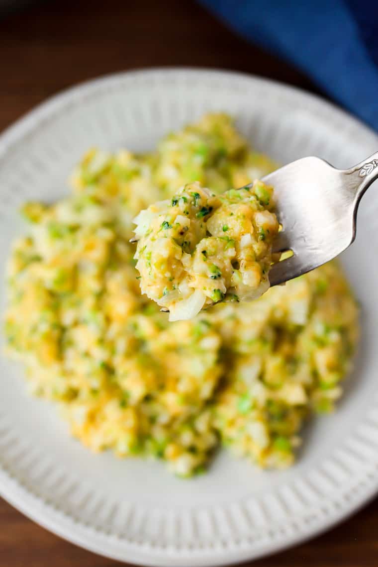 A forkful of cheesy broccoli cauliflower rice with a plate of rice and blue napkin blurred in the background