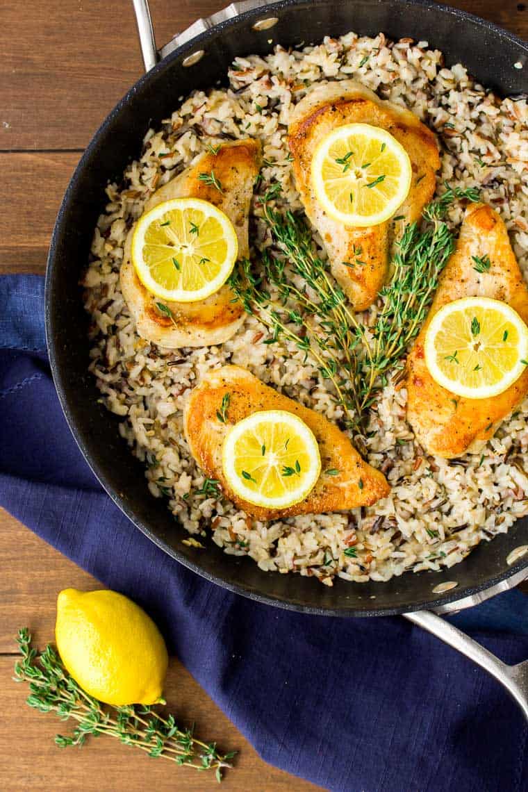 4 chicken breasts and rice topped with lemon slices and fresh thyme in a black skillet with a dark blue napkin around it and a lemon and more thyme next to it on a wood backdrop
