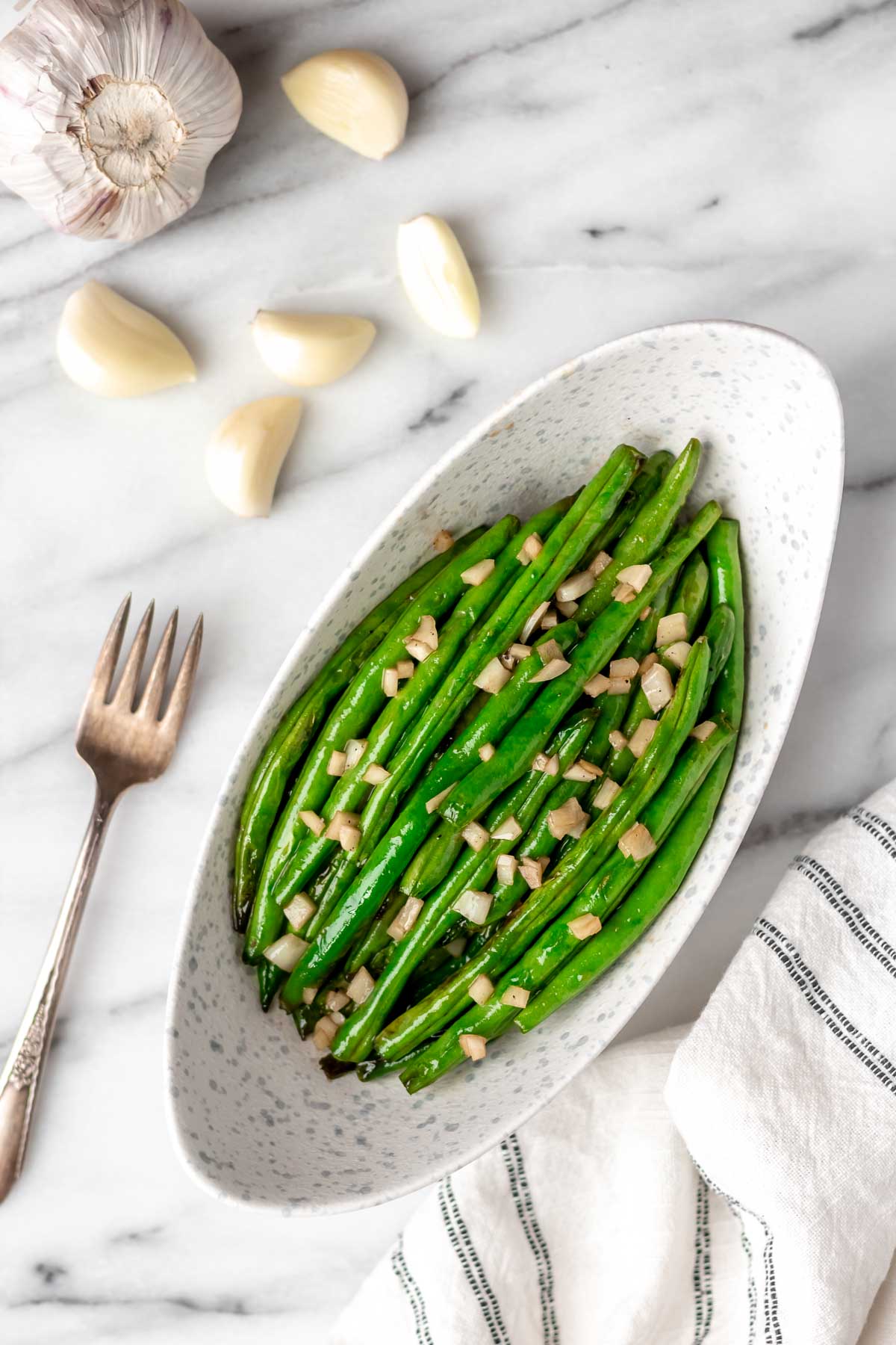 Overhead of honey garlic green beans in an oblong bowl with a fork, garlic cloves and head of garlic around it.