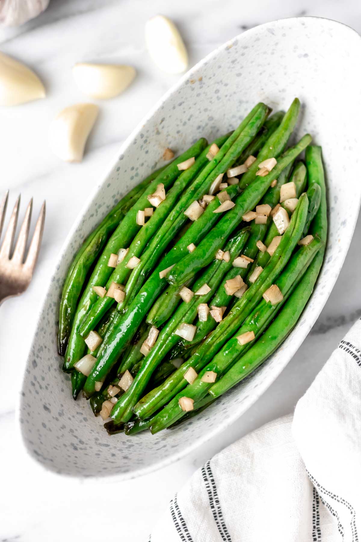 Green beans with garlic in an oblong bowl with a fork and garlic cloves around it.
