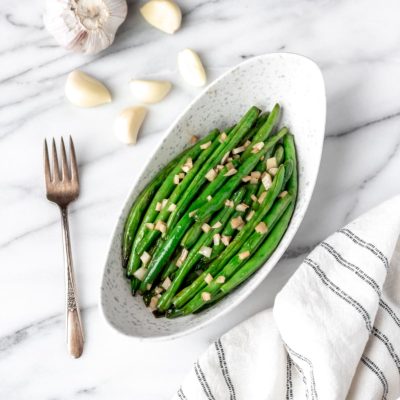 Overhead of honey garlic green beans in an oblong bowl with a fork, garlic cloves and head of garlic around it.