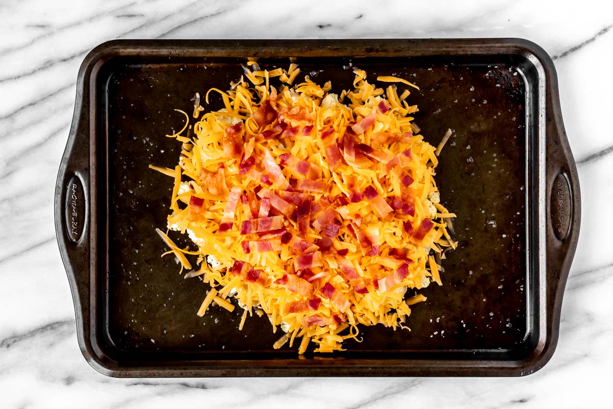 Cauliflower in a pile on a sheet pan covered in shredded cheese and pieces of bacon.