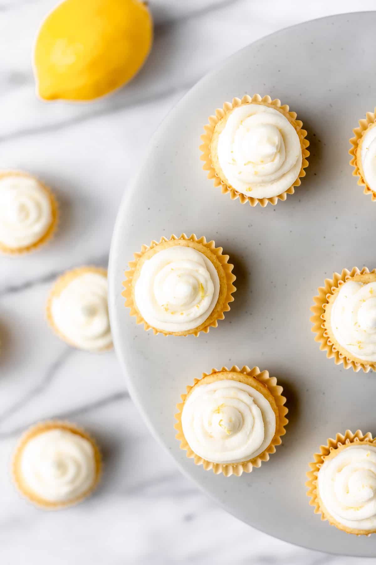 Overhead of mini lemon cupcakes on a cake stand with more on the table below with a lemon.