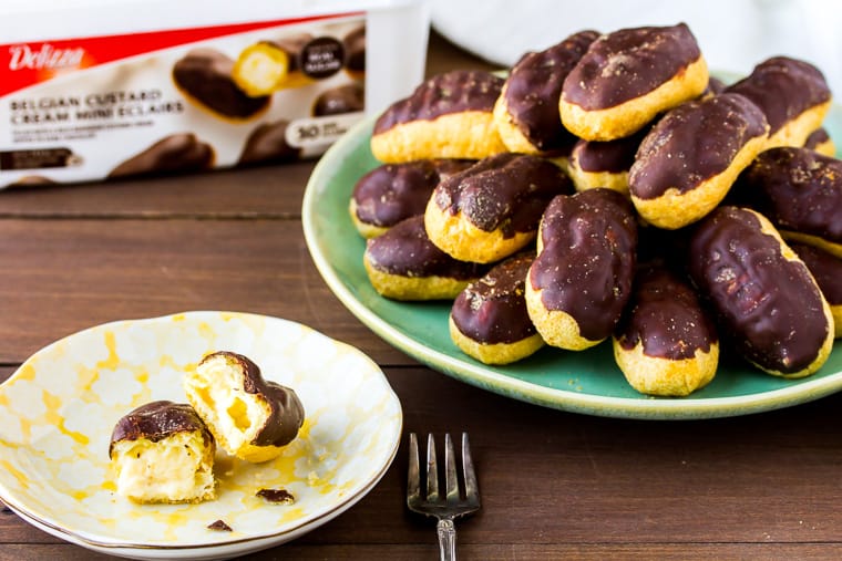 A gren serving plate full of mini chocolate eclairs with a smaller yellow plate with an eclair broken open and the box they came in in the background - all over a wood backdrop