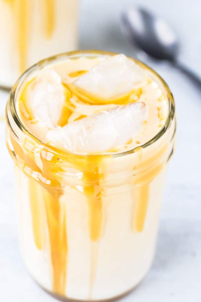 Close up of a canning jar with an ice vanilla latte topped with caramel syrup on a white table with a second jar and a spoon in the background