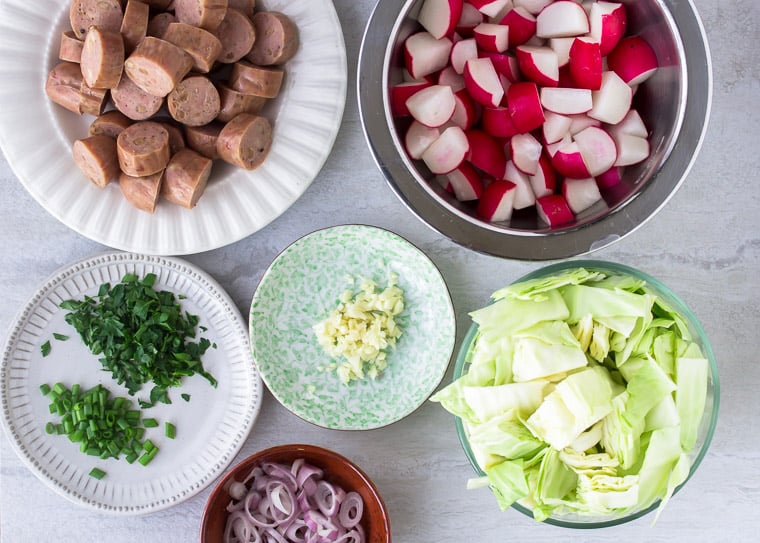 All of the prepped ingredients needed for the sausage, cabbage, and radish foil packets laid out in bowls and plates over a white backdrop