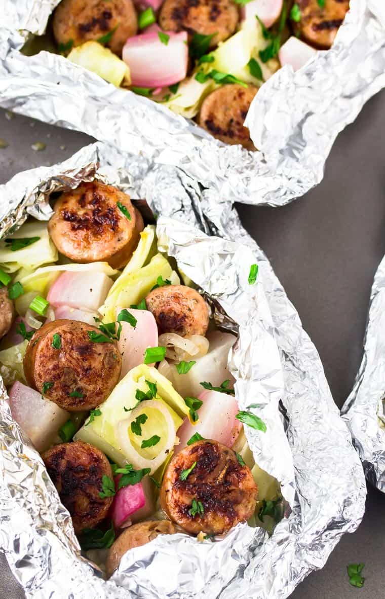 2 foil packets filled with cabbage, sausage, and radishes and topped with fresh herbs on a gray baking sheet