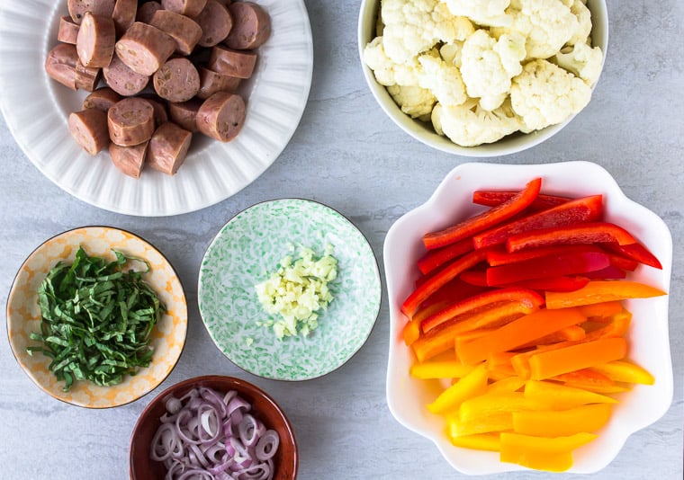 All of the ingredients needed to make sausage, pepper, and cauliflower foil packets in bowls and plates over a white background
