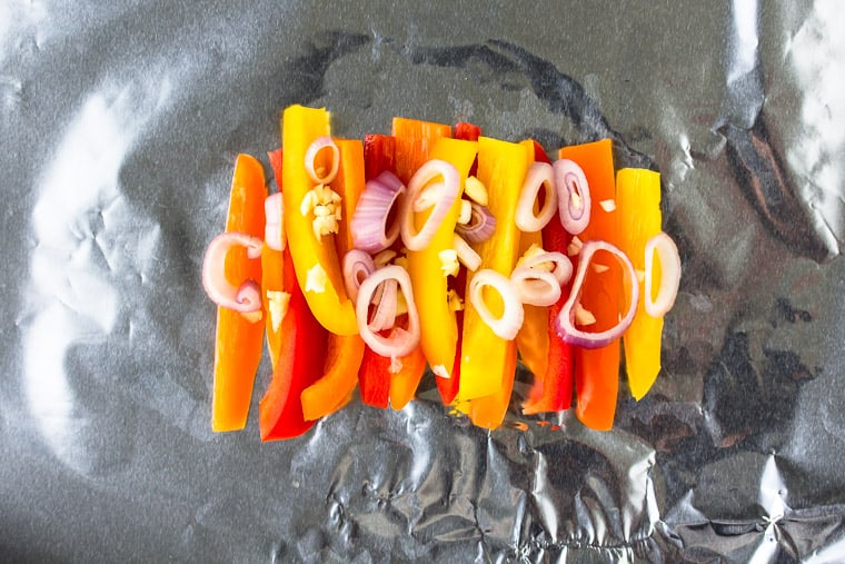 red, orange and yellow pepper strips with shallots and garlic laid out on a sheet of foil