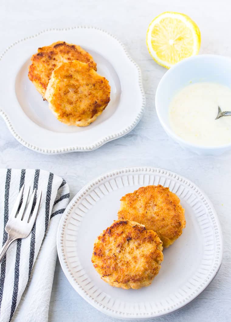 4 Keto Shrimp Cakes on 2 small white plates with a black and white striped napkin, fork, half a lemon, and a small blue bowl of aioli in the background over a white back drop