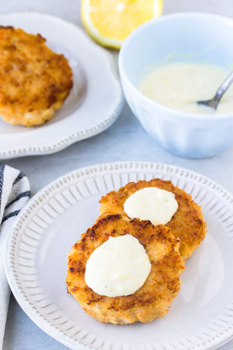 2 Keto Shrimp Cakes on a white plate with a second plate, blue bowl with aioli, a lemon, and a balck and white napkin in the background all over a white backdrop