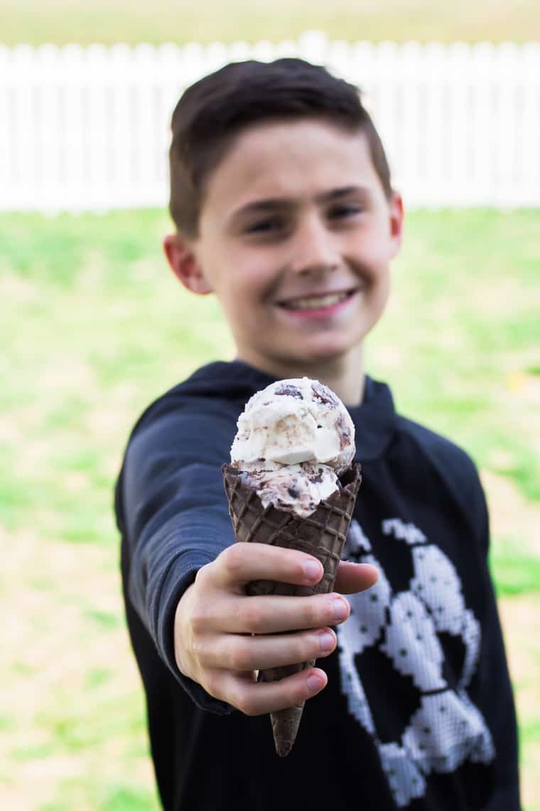 Boy holding out a chocolate ice cream cone filled with vanilla ice cream with candy in it with grass and a white picket fence in the background