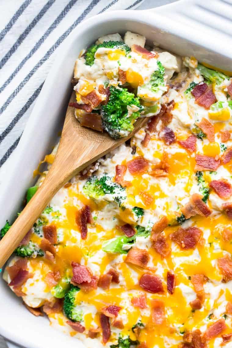 Cheesy broccoli and chicken ranch casserole in a white casserole dish with a wooden spoon taking a scoop out and a black and white striped napkin in the background