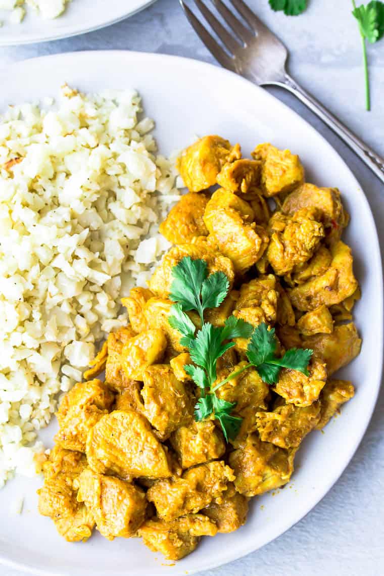 Close up of vadouvan curry chicken on a white plate with cauliflower rice and a second plate, fork, and cilantro leaves in the background. all over a white background