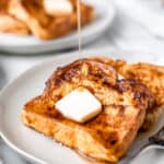 Two slices of French toast on a white plate topped with butter with syrup being drizzled on and text overlay.