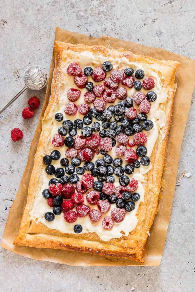 An overhead view of a whole puff pastry tart topped with cream cheese and berries on a white backdrop