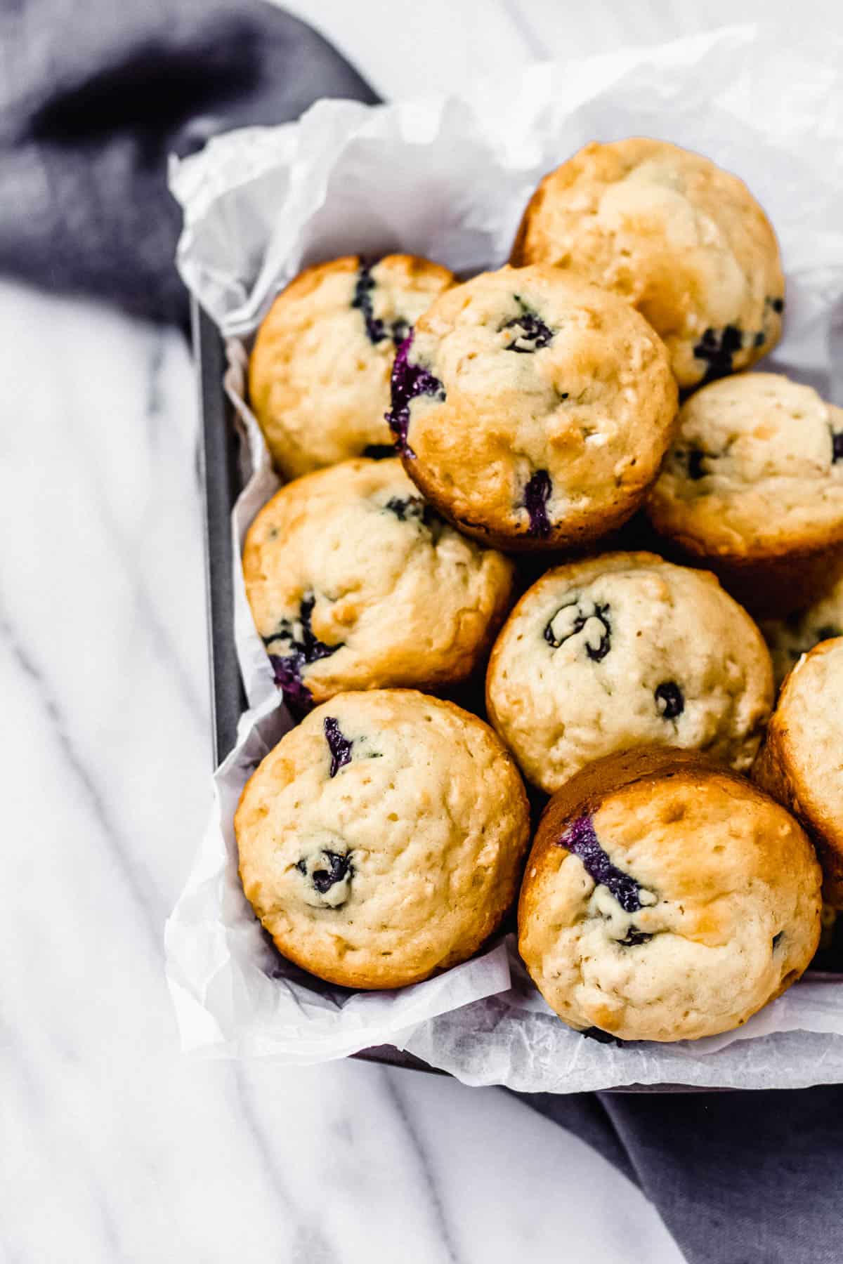 Blueberry oat muffins in a parchment paper lined tray with a gray towel under it on a marble backdrop