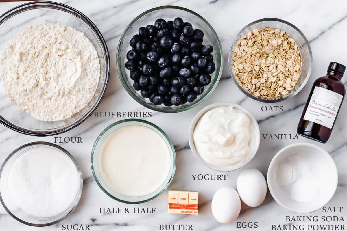 Ingredients needed to make blueberry oat muffins on a marble backdrop with labels