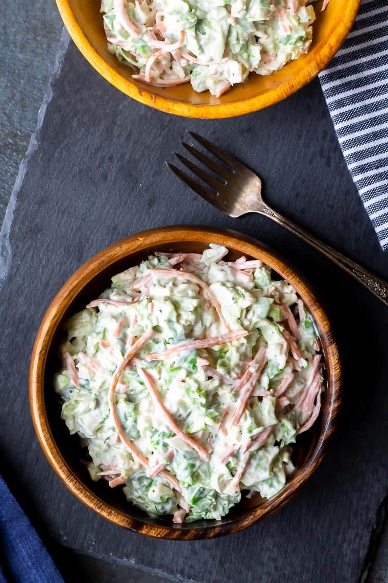 2 wooden bowls of brussels sprouts coleslaw on a piece of slaw over a gray background with a fork and blue and white striped towel