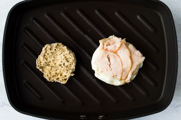 Two english muffin halves in a black grill pan. One is plain and the other has turkey and cheese on it.